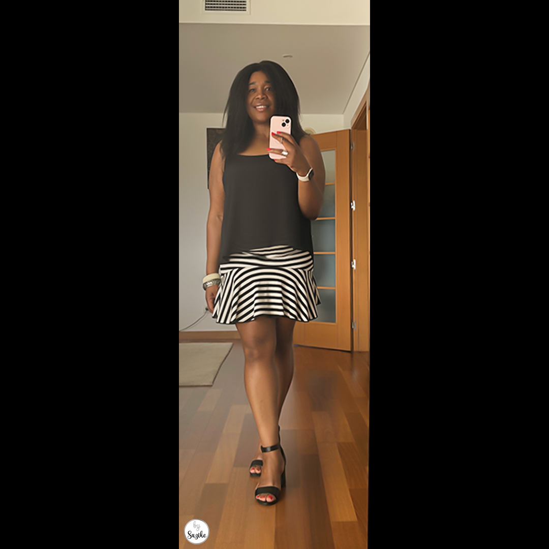 Woman posing in a black sleeveless top and striped skirt, holding her phone, wearing a trendy summer outfit with strappy black heels.
