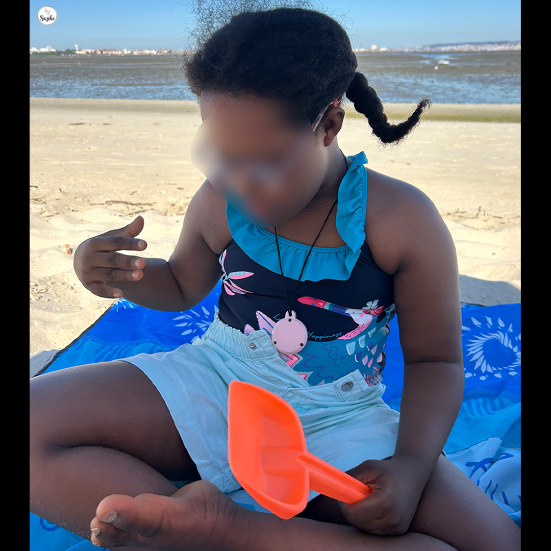 Girl playing on the sand during a beach day with kids, wearing a swimsuit and blue shorts, sitting on a blue towel.