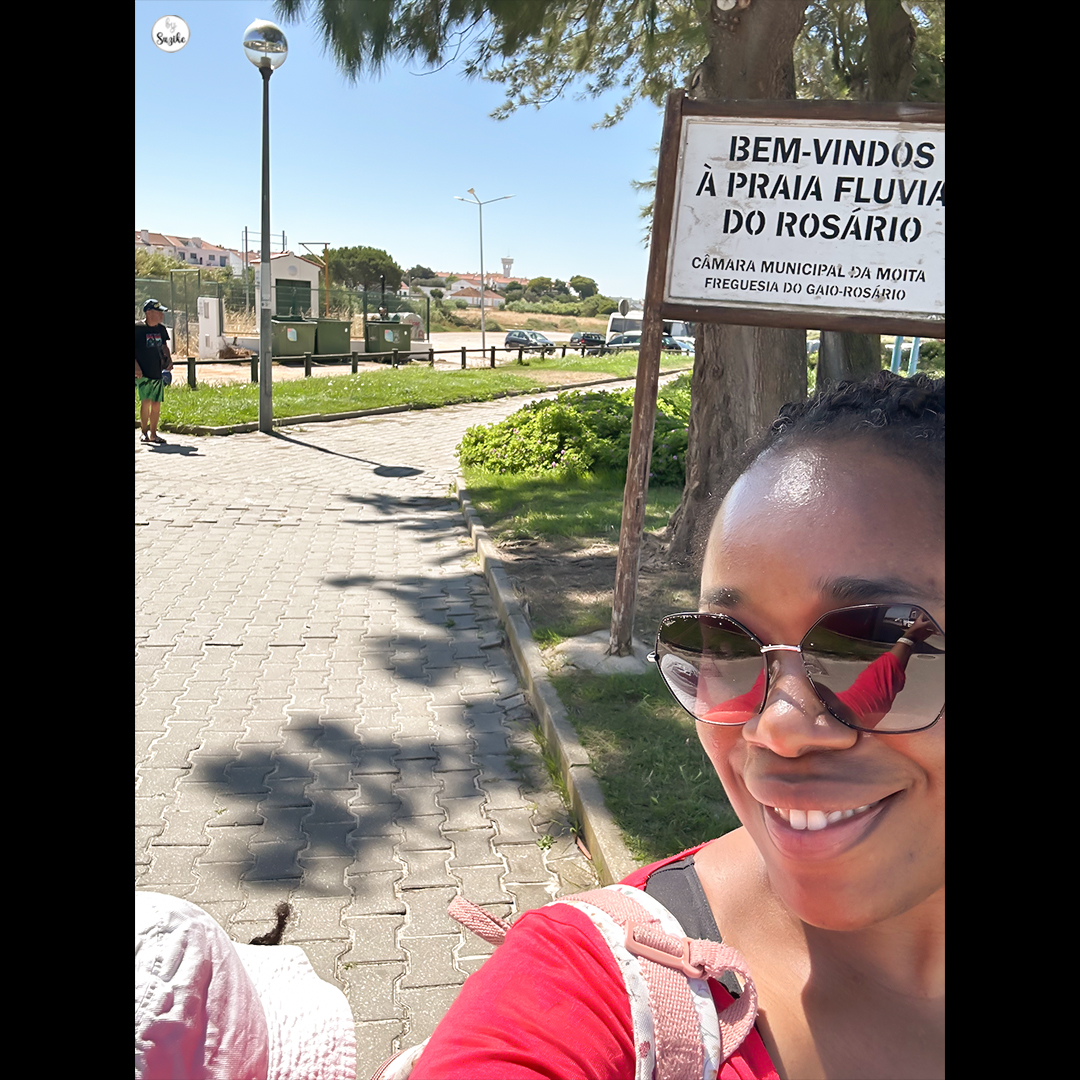 Smiling woman wearing sunglasses taking a selfie at the entrance of Praia Fluvial do Rosário, river beach in Moita, Portugal. Welcome sign and sunny summer day visible in the background.
