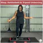 Suzike standing on her new red and black step during a home gym unboxing, smiling and showing the new workout equipment — step, kettlebell, and tripod.