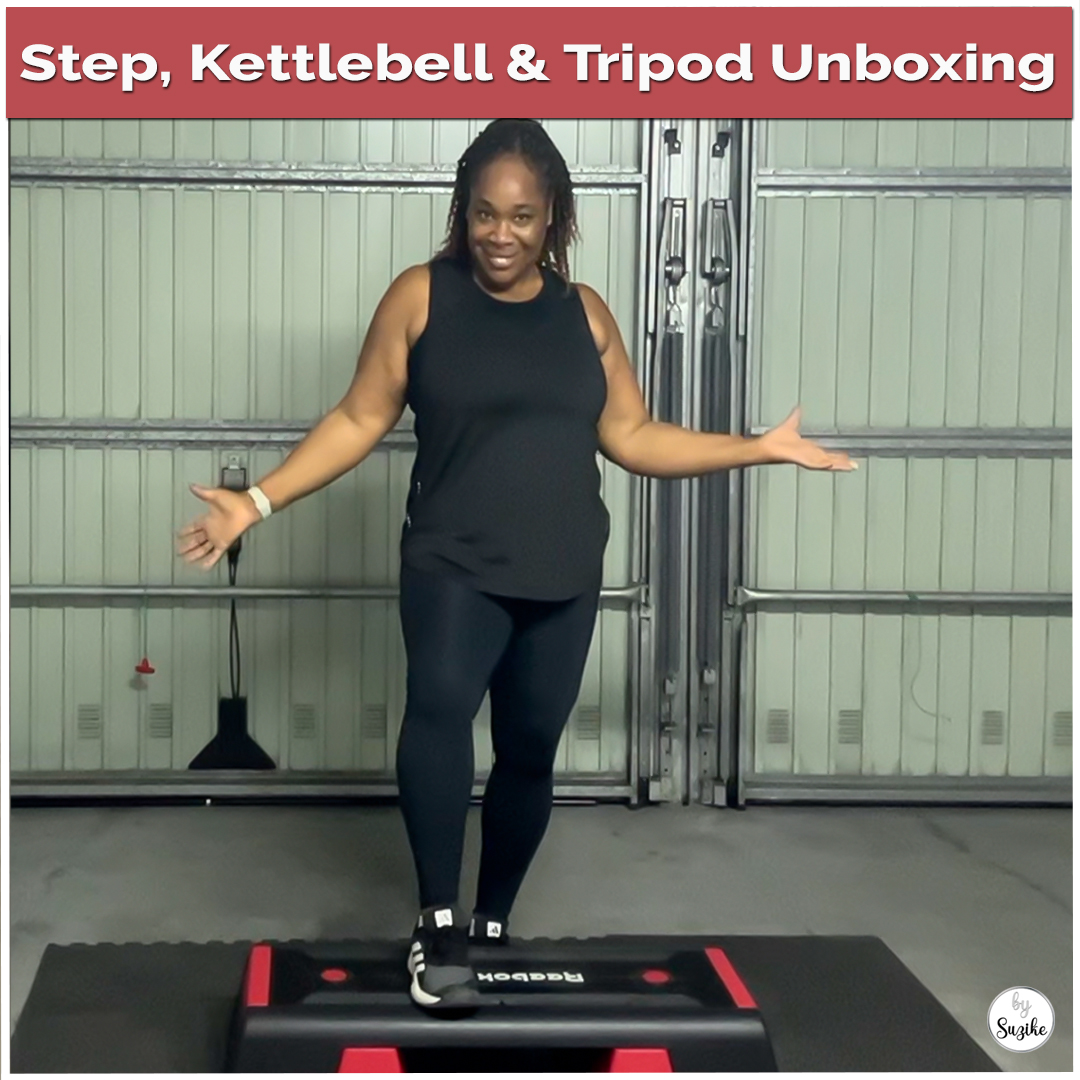 Suzike standing on her new red and black step during a home gym unboxing, smiling and showing the new workout equipment — step, kettlebell, and tripod.