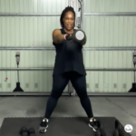 Woman training in a home gym with a kettlebell, mid-workout stance, focused expression, indoor lighting, minimal background.