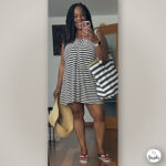 Mirror selfie of Suzike holding a summer hat and wearing a casual beach striped dress with a large striped tote bag.