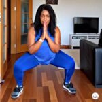 Woman doing a deep sumo squat at home with hands together, wearing a blue workout outfit and trainers.