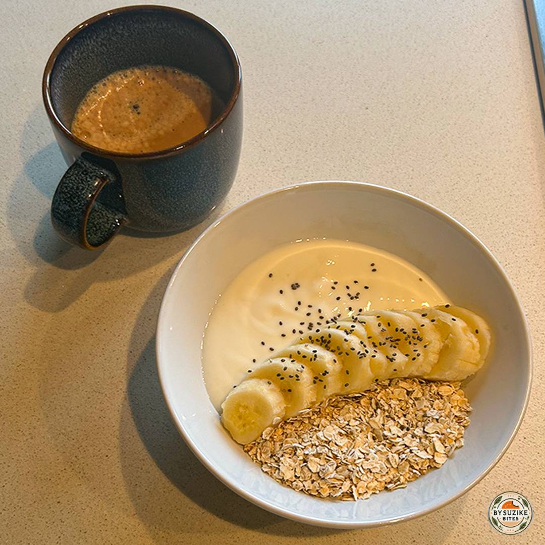 Bowl with yogurt, rolled oats, and sliced banana served with a cup of coffee on a kitchen counter.