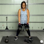 20 minute kettlebell leg workout at home with woman holding two kettlebells on a mat, dumbbells on the floor, and a home gym garage background.