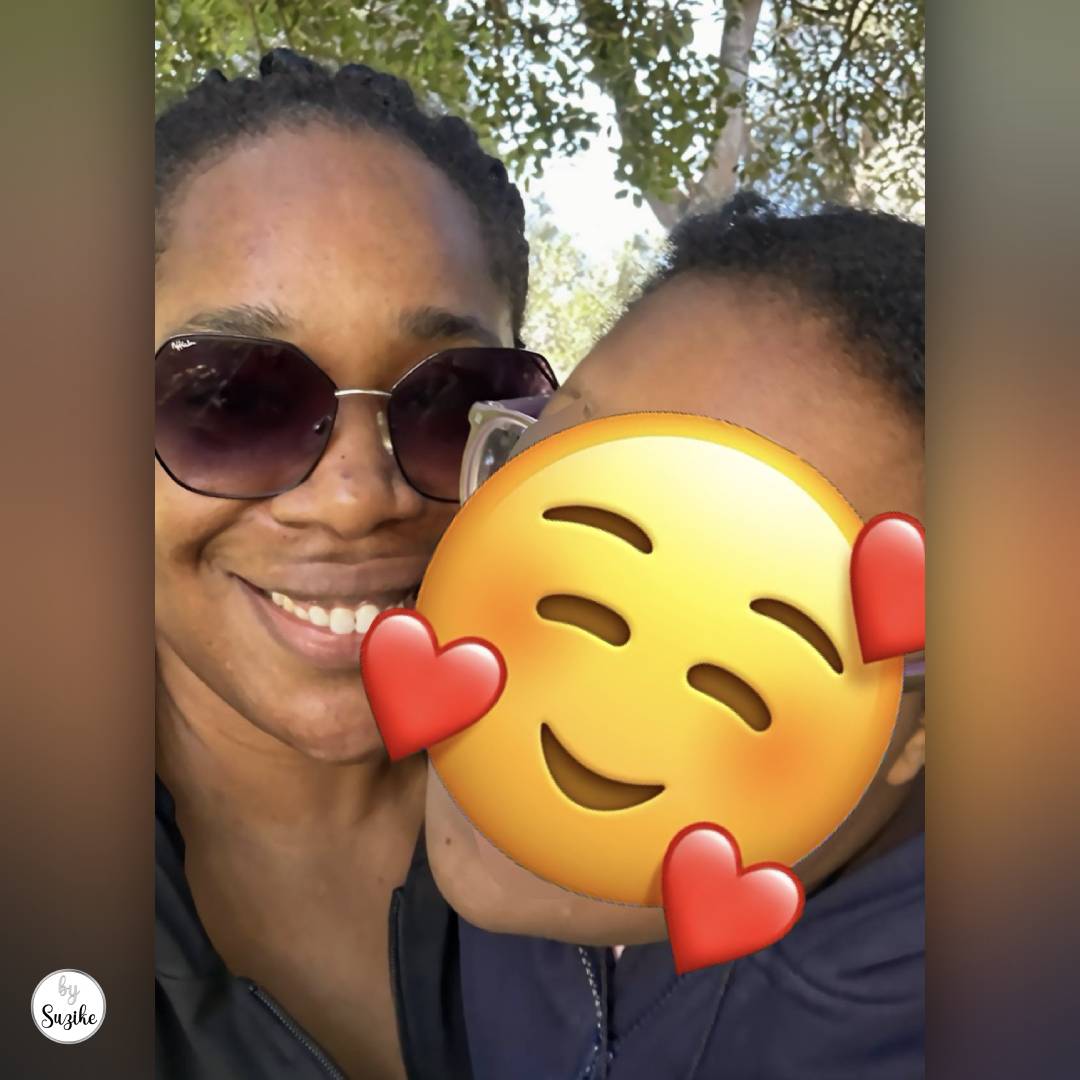 Close-up selfie of a smiling mother wearing sunglasses with her child during a sunny weekend walk in Lisbon; child’s face covered with an emoji for privacy.