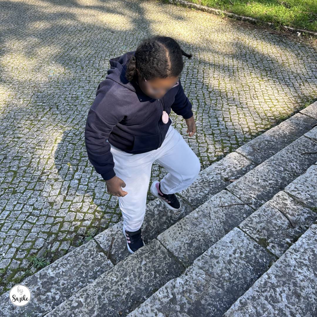 Child climbing up outdoor stone stairs during a sunny walk, wearing a navy hoodie, light joggers, and trainers; cobblestone path in the background, face blurred for privacy.