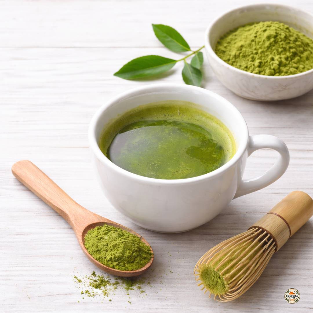 Close up of a cup of matcha tea with matcha powder, a bamboo whisk and green tea leaves on a light wooden surface.