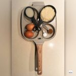 Overhead photo of a square nonstick egg pan on a kitchen counter with two eggs, measuring cups, and ingredients inside the pan.