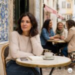 Woman over 40 sitting alone at an outdoor café in Portugal, with other women chatting in the background, for an article about how to make friends in Portugal after 40.