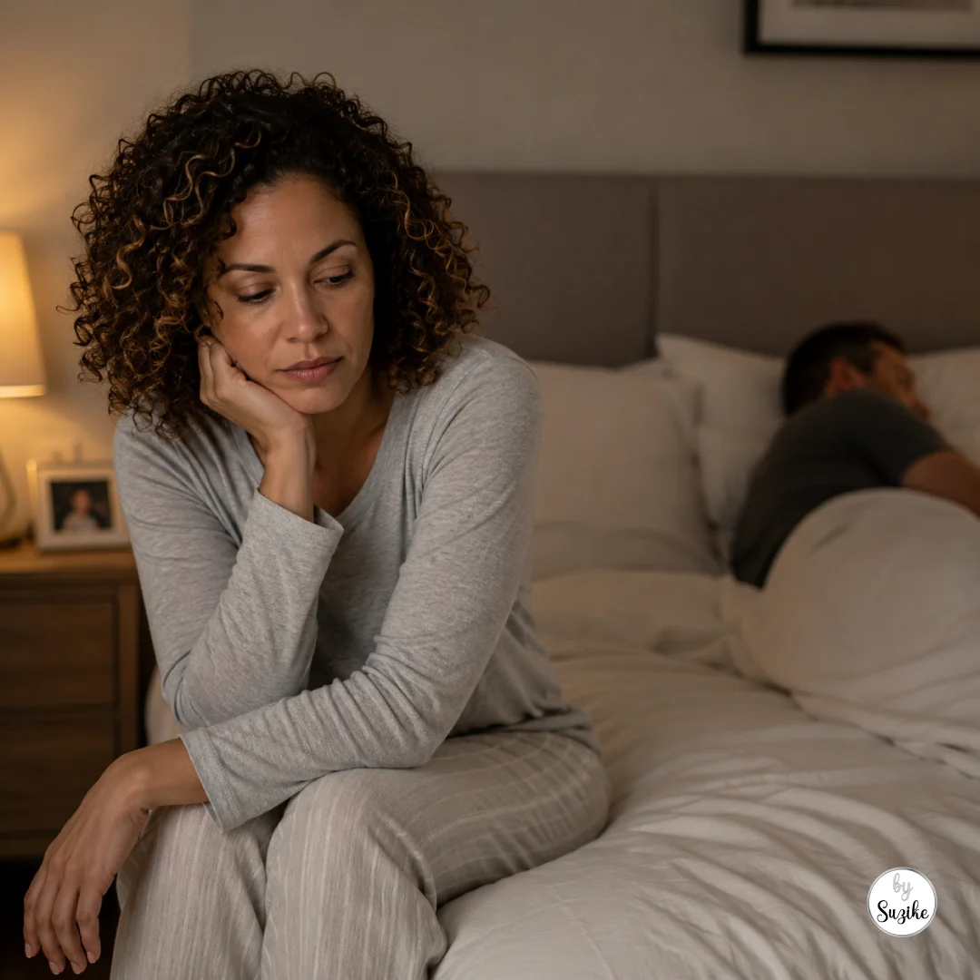 low-libido-after-40-bedroom-distance-couple Woman sitting on the edge of a bed looking tired while her partner lies in the background, illustrating low libido after 40 and emotional distance in a long-term relationship.